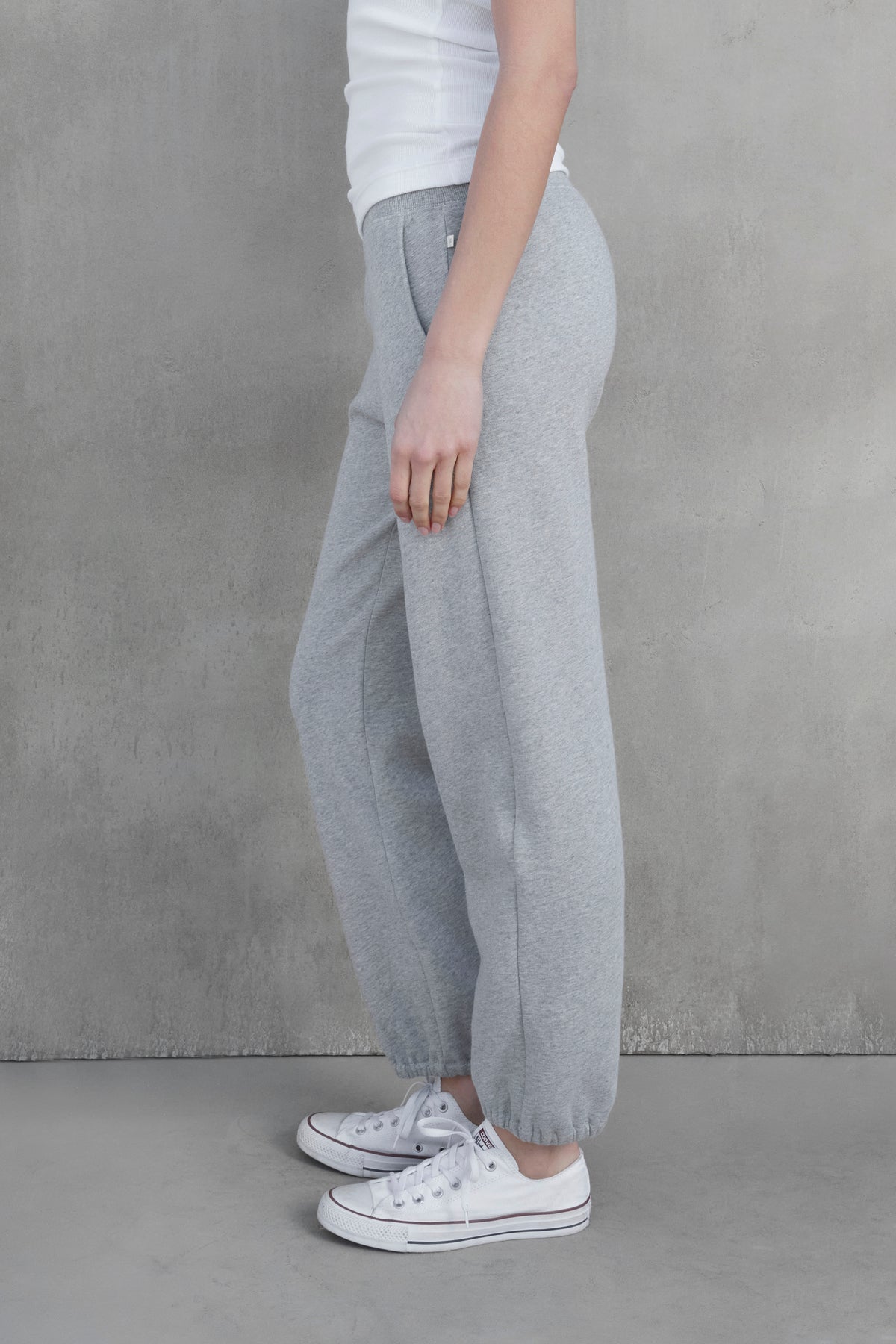   A person in a white shirt and the ZUMA SWEATPANT by Velvet by Jenny Graham stands sideways against a gray concrete wall, wearing white sneakers. 