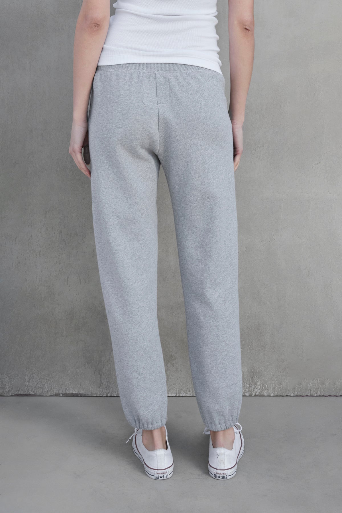 Person standing against a grey wall, wearing ZUMA SWEATPANT by Velvet by Jenny Graham in grey organic cotton, a white sleeveless shirt, and white sneakers. The person is viewed from behind.-37659947860161