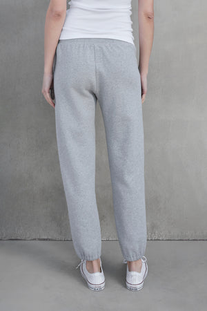 Person standing against a grey wall, wearing ZUMA SWEATPANT by Velvet by Jenny Graham in grey organic cotton, a white sleeveless shirt, and white sneakers. The person is viewed from behind.