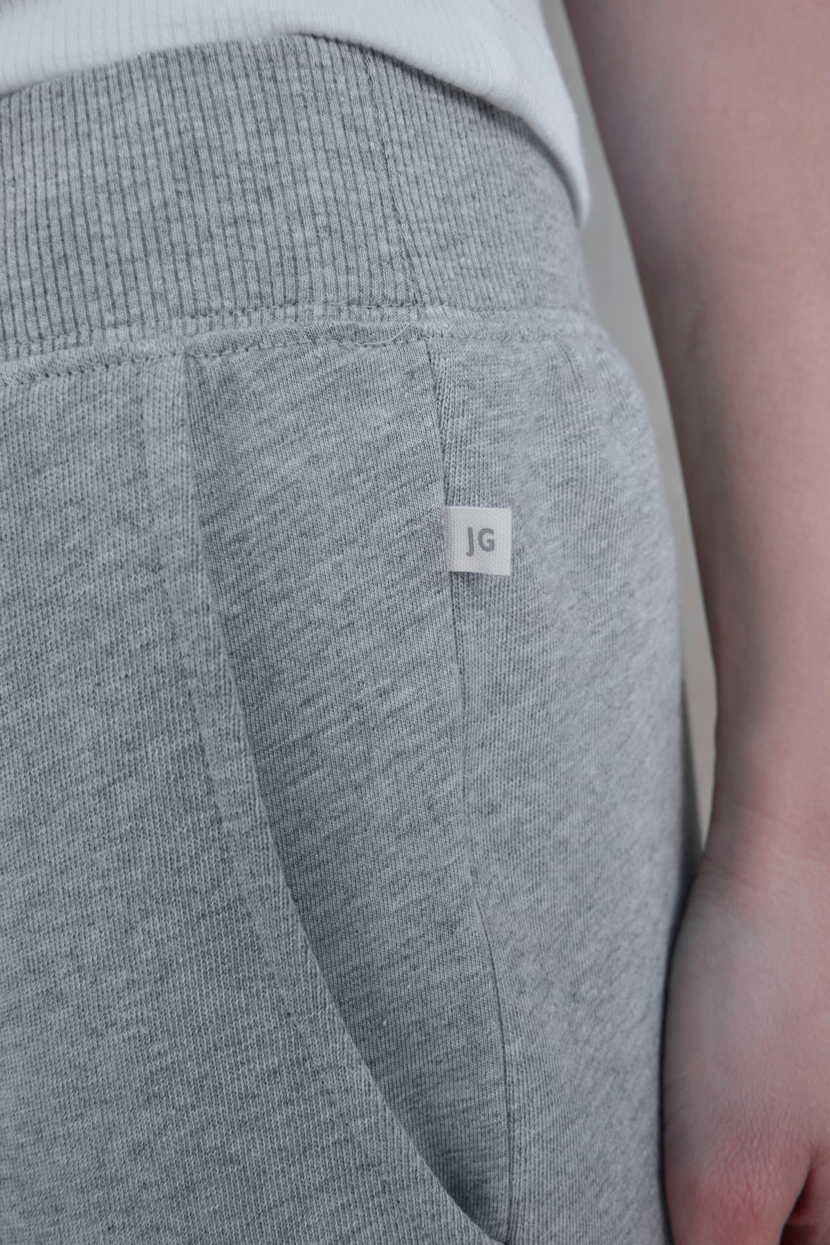   Close-up of a person wearing Velvet by Jenny Graham's ZUMA SWEATPANT, crafted from organic fleece in a light gray shade. The joggers feature a small tag with the initials 