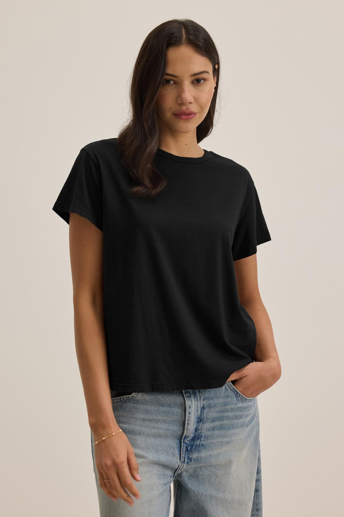 A woman with long dark hair wears the TOPANGA TEE from Velvet by Jenny Graham, a relaxed-fit classic black t-shirt, paired with organic cotton jeans. She strikes a pose with one hand in her pocket against a simple, eco-friendly backdrop.-37817545162945