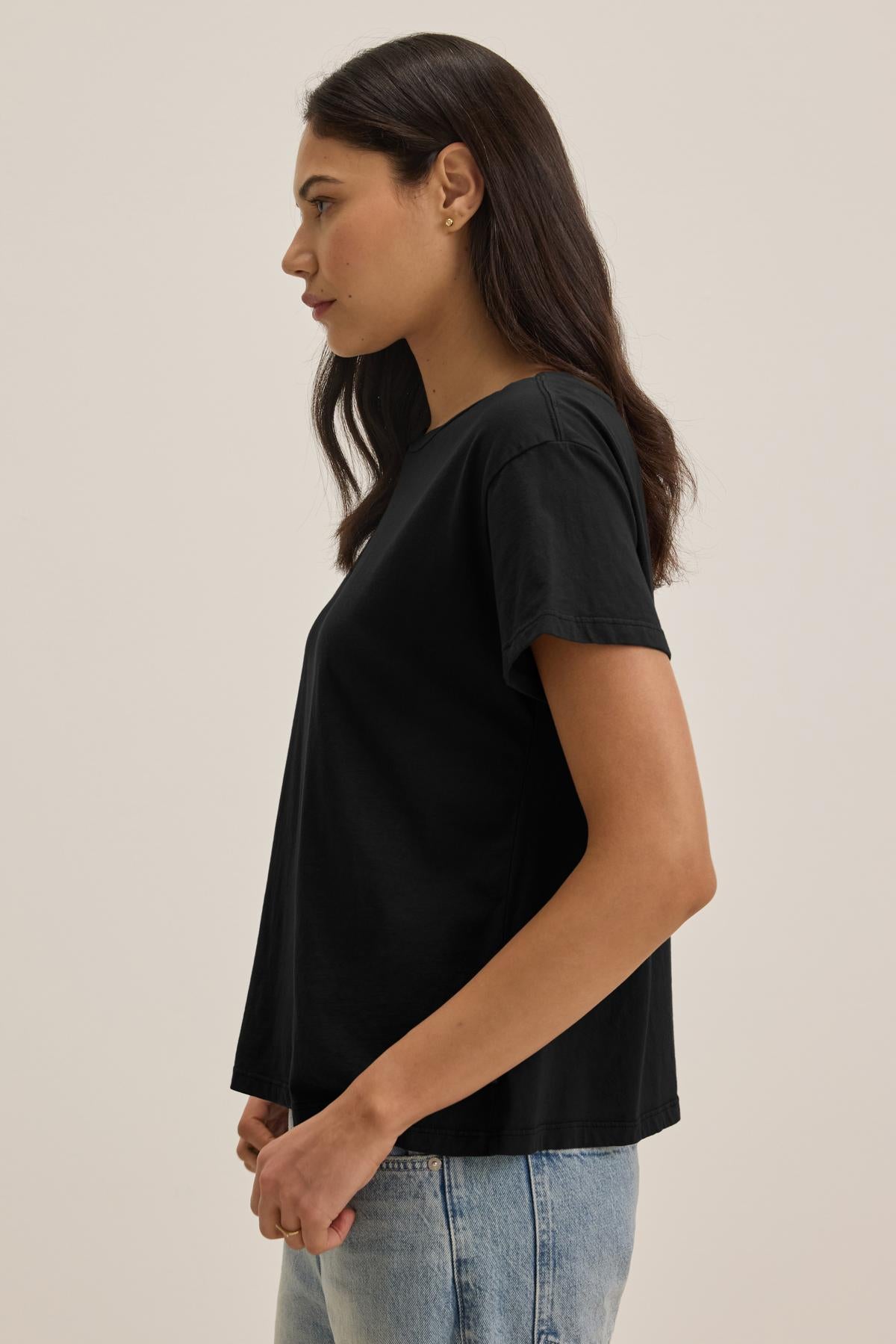   Profile view of a woman with long dark hair wearing the TOPANGA TEE from Velvet by Jenny Graham, a relaxed-fit classic black t-shirt, and light blue jeans made from organic cotton. She is standing against a plain background. 