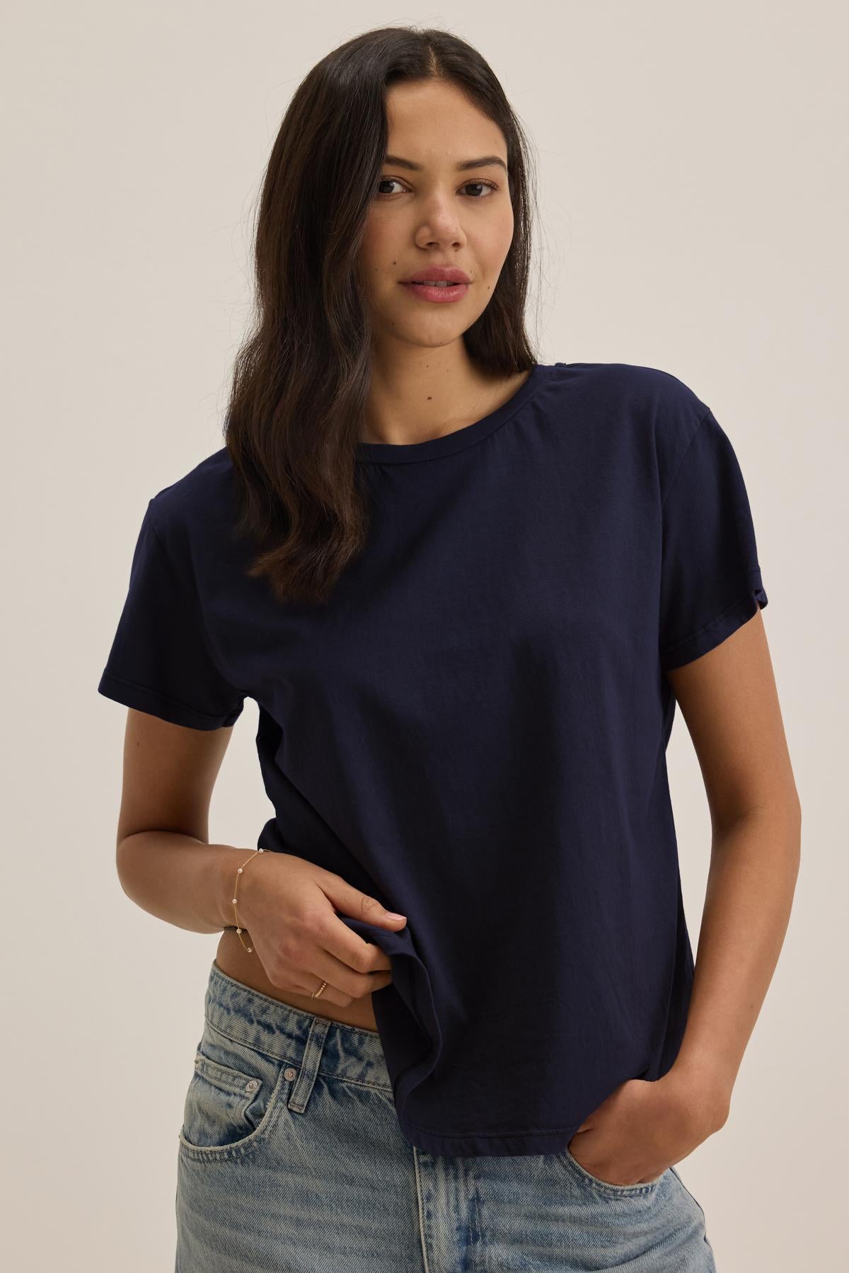   A person with long dark hair wearing the TOPANGA TEE, a navy blue, relaxed-fit, short-sleeved shirt by Velvet by Jenny Graham, and light blue jeans stands against a plain background, looking straight ahead with one hand in their pocket. 