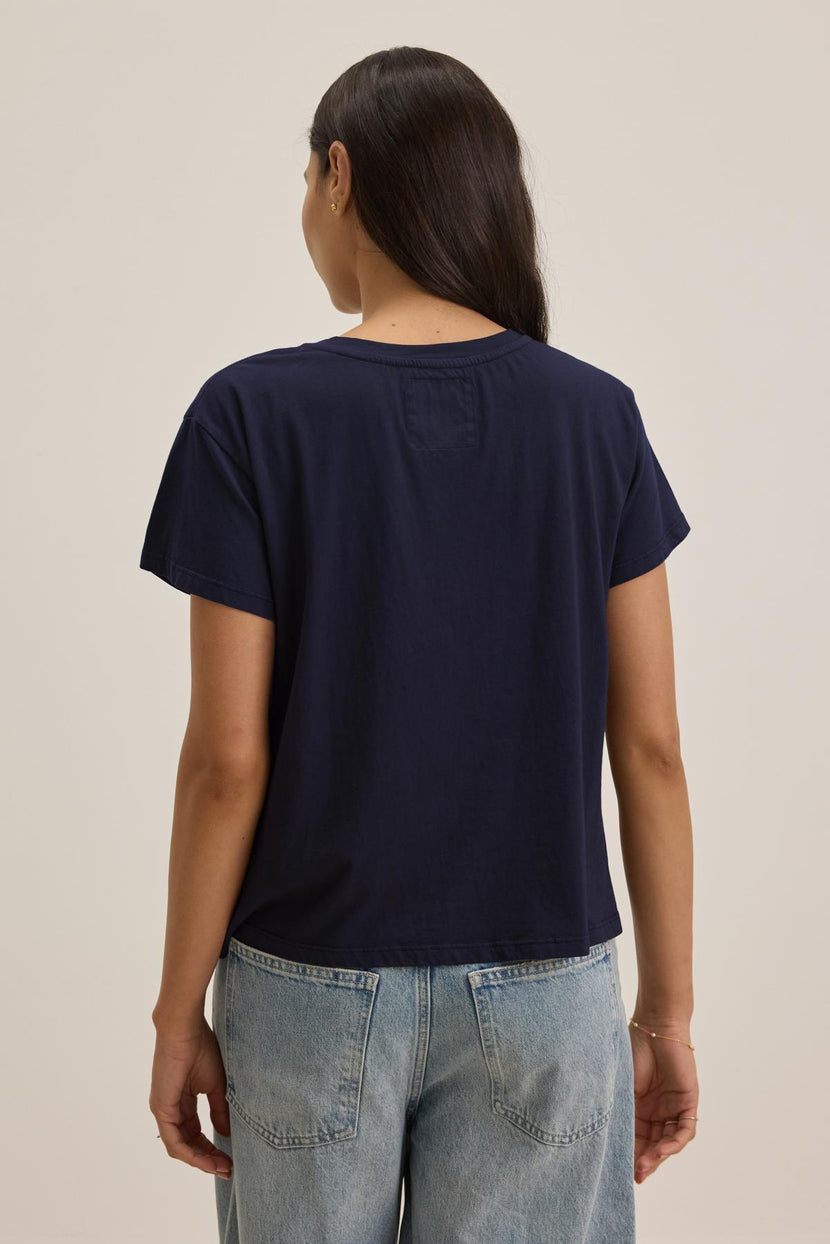 A person with long dark hair wearing a navy blue, relaxed-fit TOPANGA TEE made of organic cotton from Velvet by Jenny Graham and light blue jeans is seen from the back, standing against a plain background.