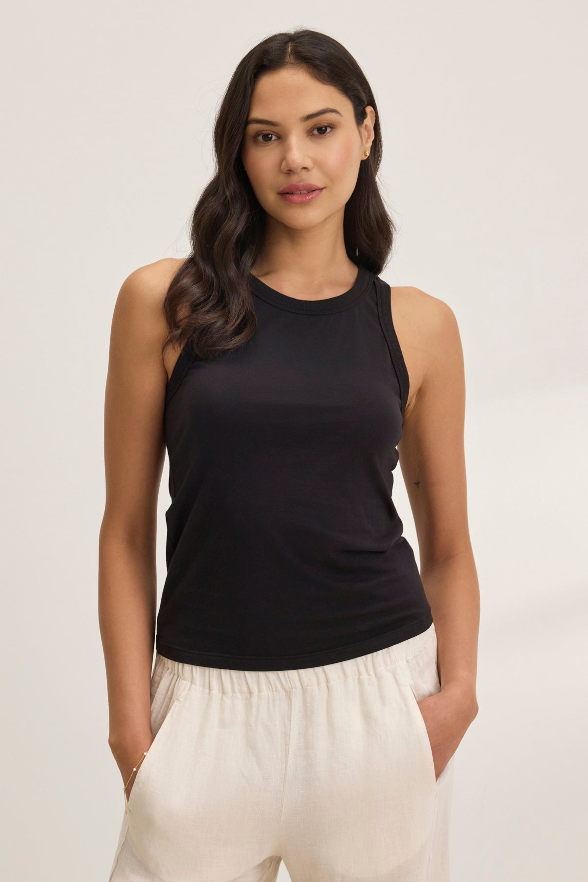   A person stands against a plain background, wearing the GREER TANK TOP by Velvet by Graham & Spencer, a fitted black tank paired with light-colored organic cotton jersey pants. 