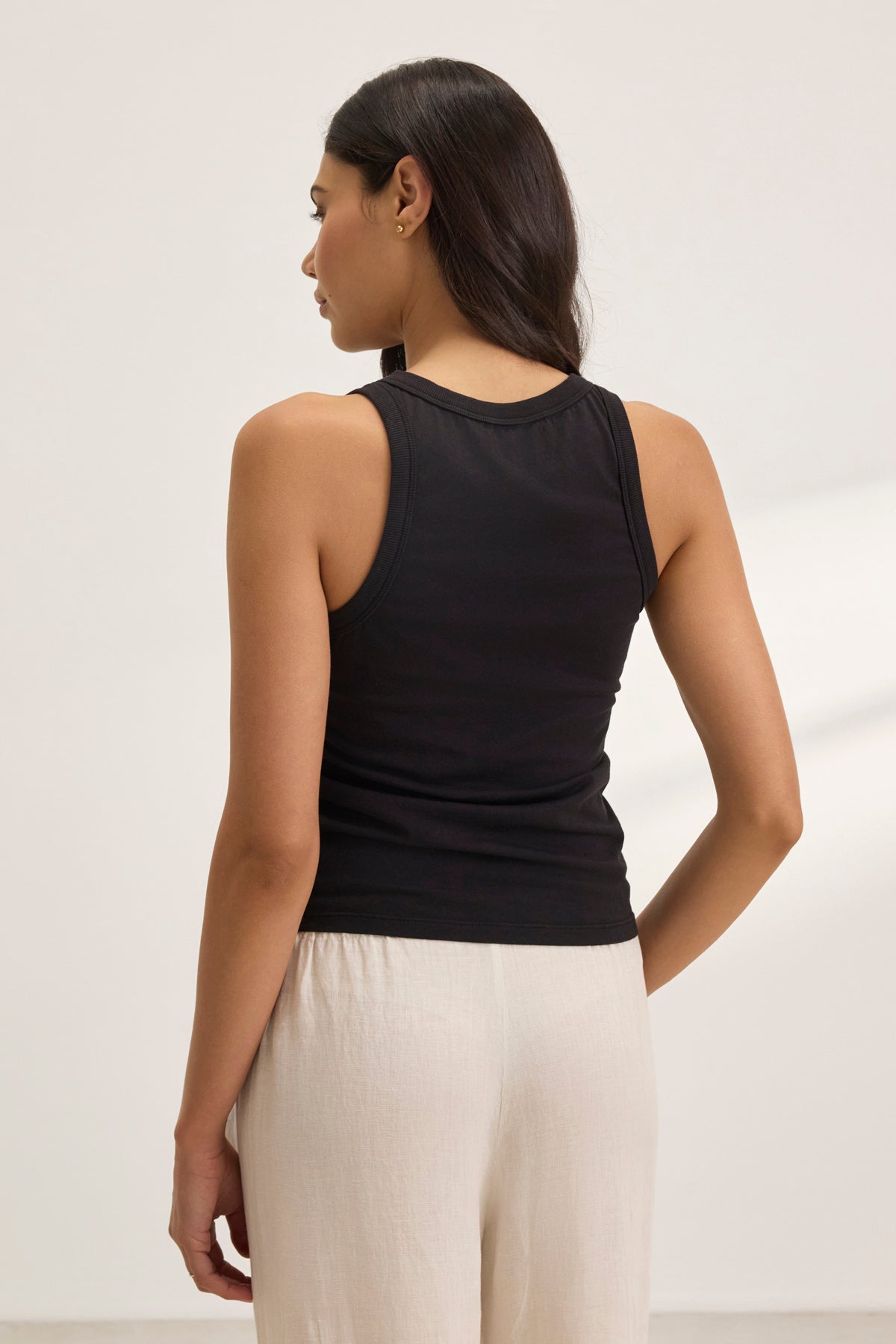   Someone with long hair, viewed from behind, wears a GREER TANK TOP by Velvet by Graham & Spencer made of organic cotton jersey and light-colored pants against a plain background. 