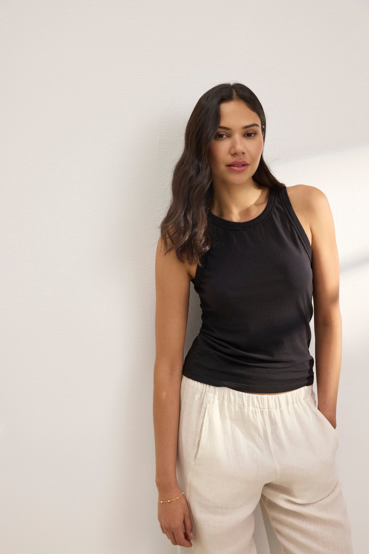   A woman with long hair in a GREER TANK TOP by Velvet by Graham & Spencer, made of organic cotton jersey, and wearing light pants leans gracefully against a white wall. 