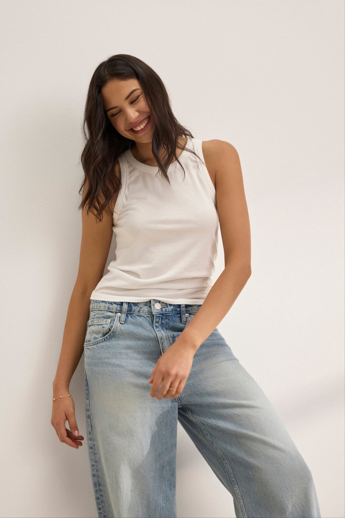   A woman wears the GREER TANK TOP by Velvet by Graham & Spencer, a white fitted tank made from soft organic cotton jersey, paired with blue jeans. She stands against a white background, smiling with her head tilted slightly. 