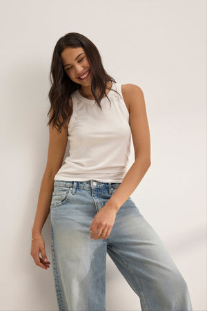 A woman wears the GREER TANK TOP by Velvet by Graham & Spencer, a white fitted tank made from soft organic cotton jersey, paired with blue jeans. She stands against a white background, smiling with her head tilted slightly.