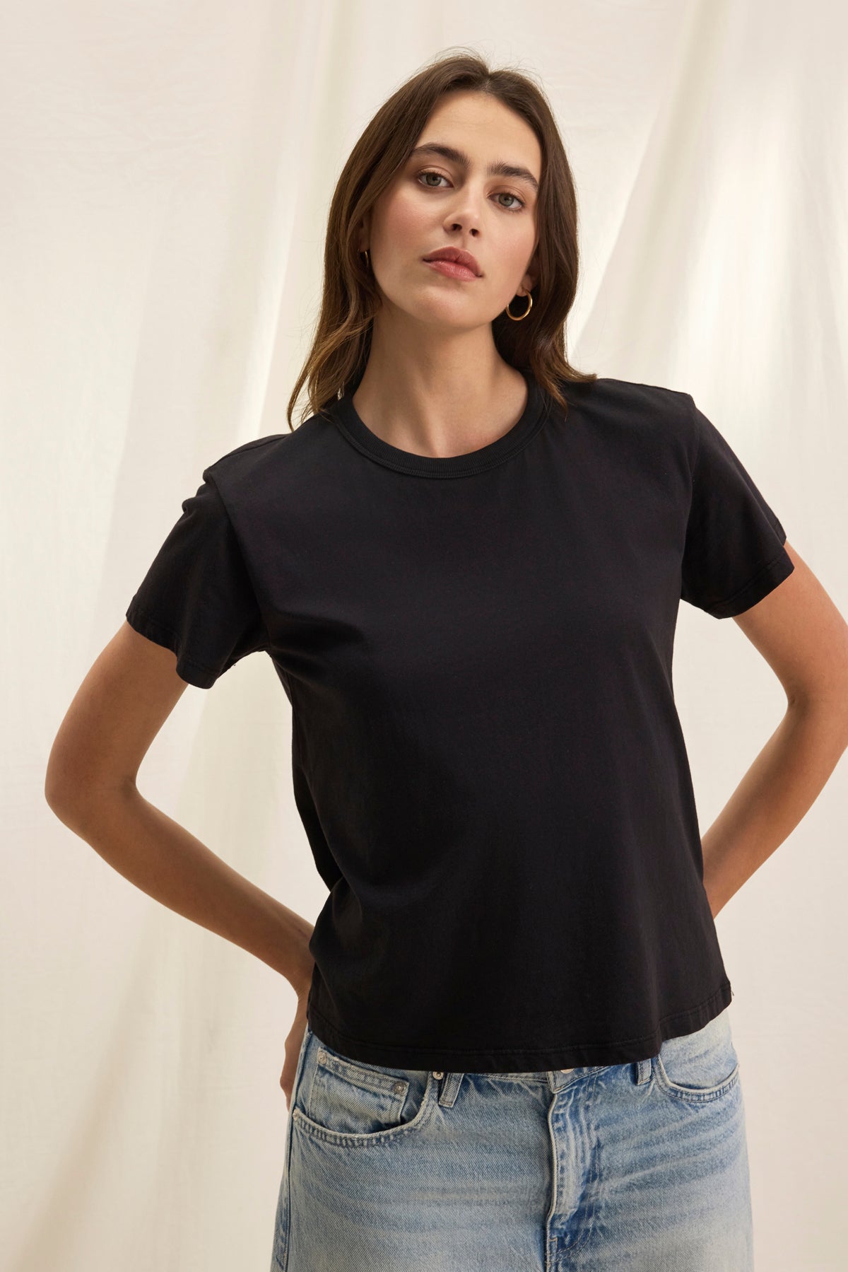 A person in a JAZZ TEE from Velvet by Graham & Spencer, featuring a crew neckline, paired with blue jeans, stands against a light backdrop with hands on hips.-38819699654849