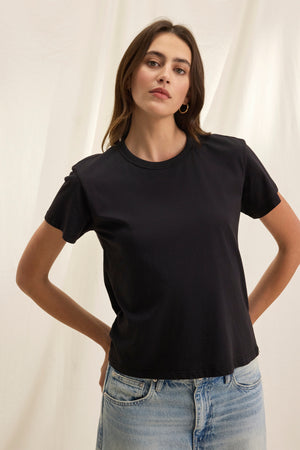 A person in a JAZZ TEE from Velvet by Graham & Spencer, featuring a crew neckline, paired with blue jeans, stands against a light backdrop with hands on hips.