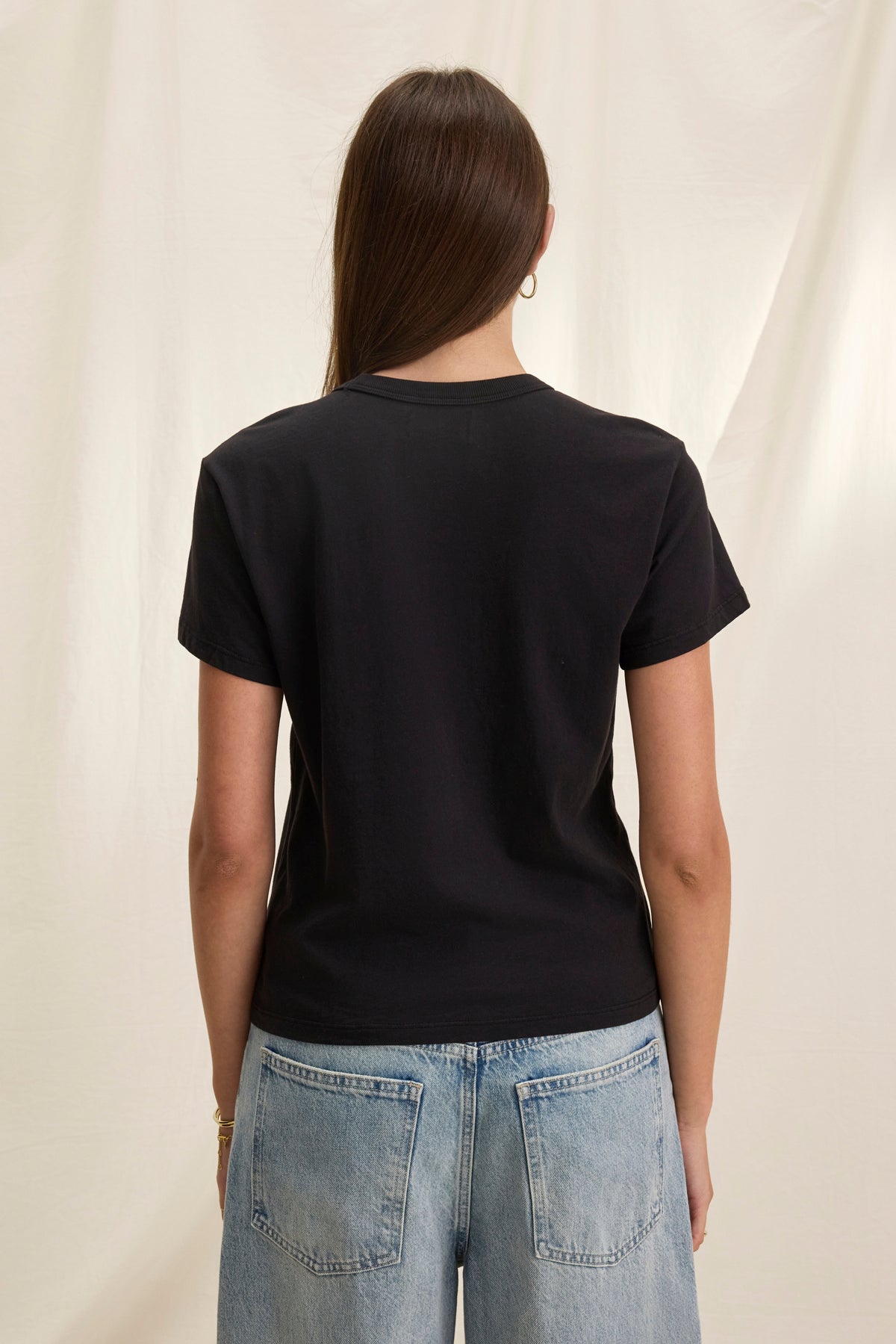 A person with long hair stands back to the camera, wearing a Velvet by Graham & Spencer JAZZ TEE in black. Made from organic cotton jersey, its relaxed boxy fit effortlessly pairs with light blue jeans for a casual yet stylish vibe.-38819699622081