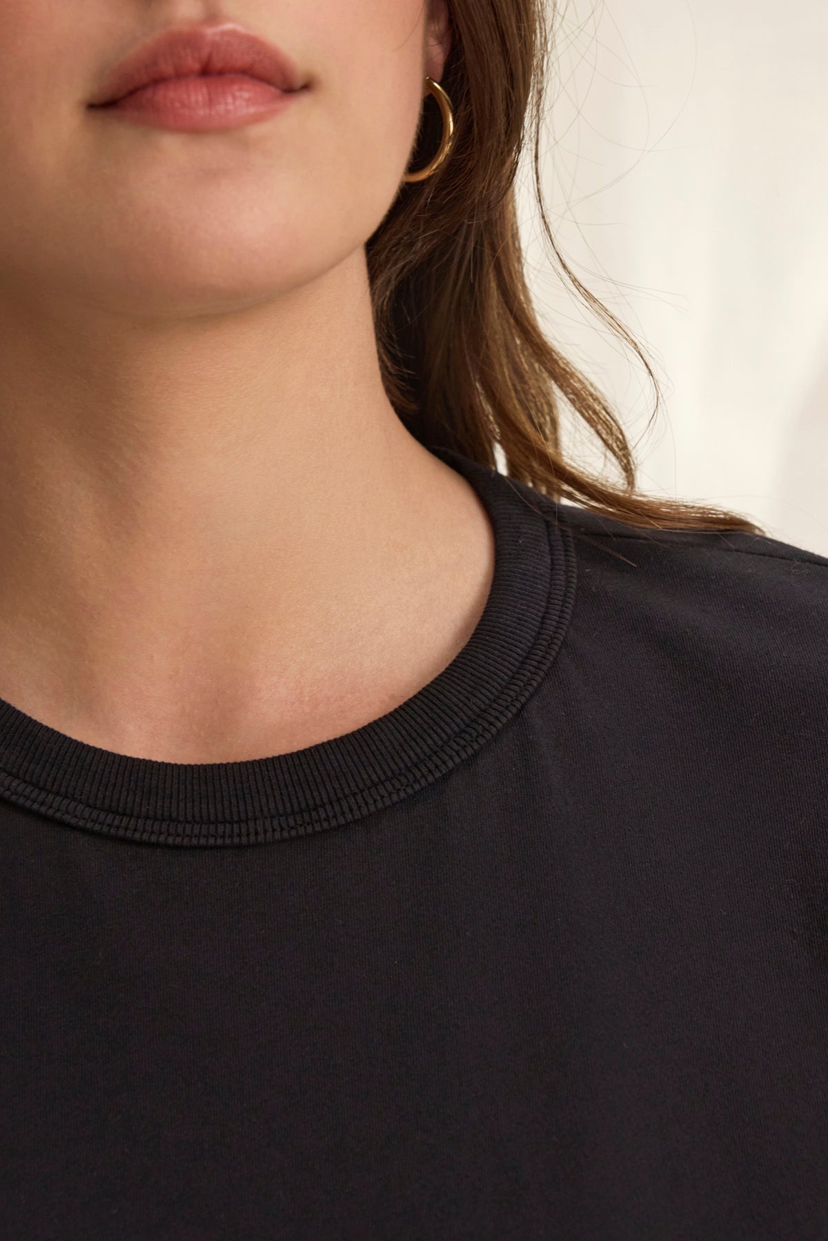   Close-up of a person wearing the JAZZ TEE by Velvet by Graham & Spencer, a black crew-neck shirt in organic cotton jersey, paired with a gold hoop earring. The relaxed silhouette is enhanced by the blurred background. 