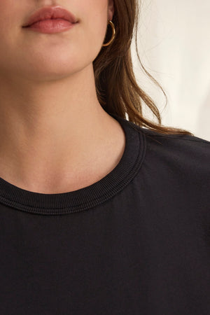 Close-up of a person wearing the JAZZ TEE by Velvet by Graham & Spencer, a black crew-neck shirt in organic cotton jersey, paired with a gold hoop earring. The relaxed silhouette is enhanced by the blurred background.