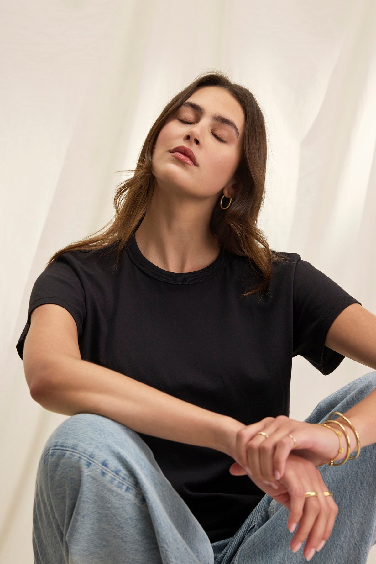 A person sits with eyes closed, surrounded by a white background, wearing Velvet by Graham & Spencer's JAZZ TEE, a black relaxed boxy silhouette t-shirt in organic cotton jersey, paired with jeans.-38819699884225