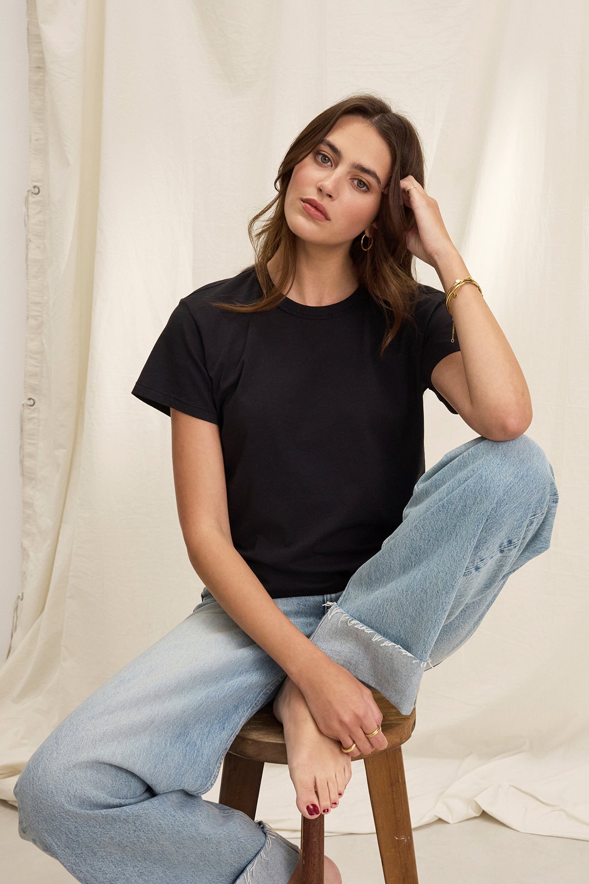   A woman in a JAZZ TEE by Velvet by Graham & Spencer sits on a stool, her elbow resting on her knee. Her long hair cascades past the black organic cotton jersey with its relaxed, boxy silhouette and crew neckline, as she wears a bracelet against the cream-colored background. 