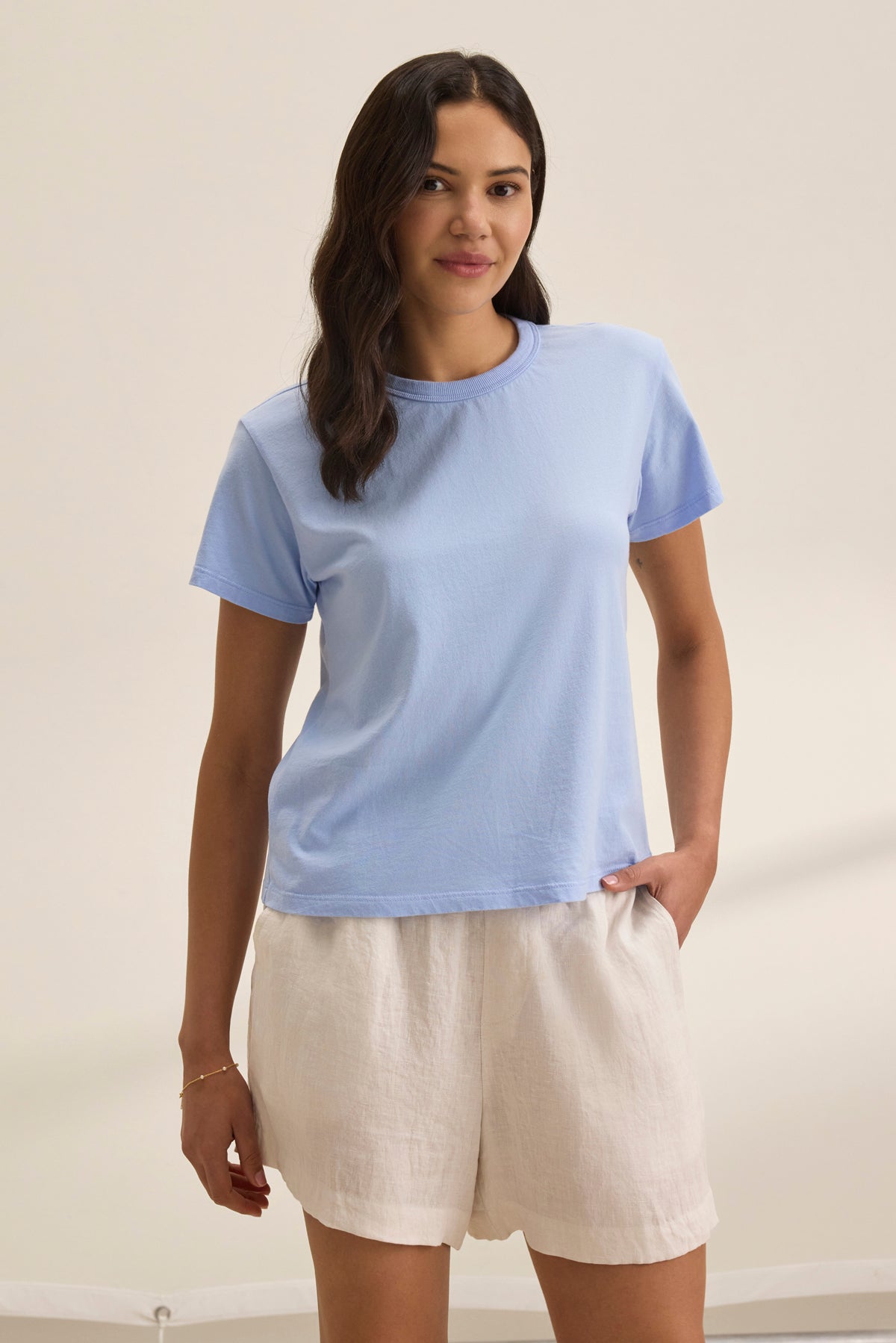 A person with long hair stands against a plain background, wearing the light blue JAZZ TEE by Velvet by Graham & Spencer, paired with white shorts.-38819699032257