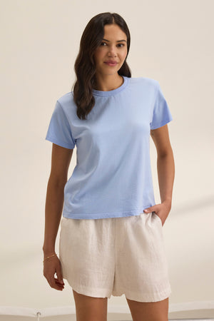 A person with long hair stands against a plain background, wearing the light blue JAZZ TEE by Velvet by Graham & Spencer, paired with white shorts.