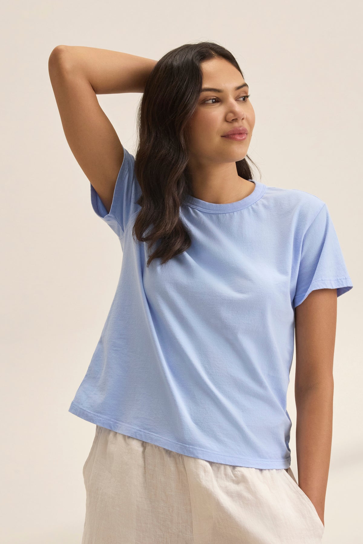 Person wearing a JAZZ TEE by Velvet by Graham & Spencer in light blue organic cotton jersey and relaxed beige pants, posing with one arm raised and looking to the side against a neutral background.-38819699785921