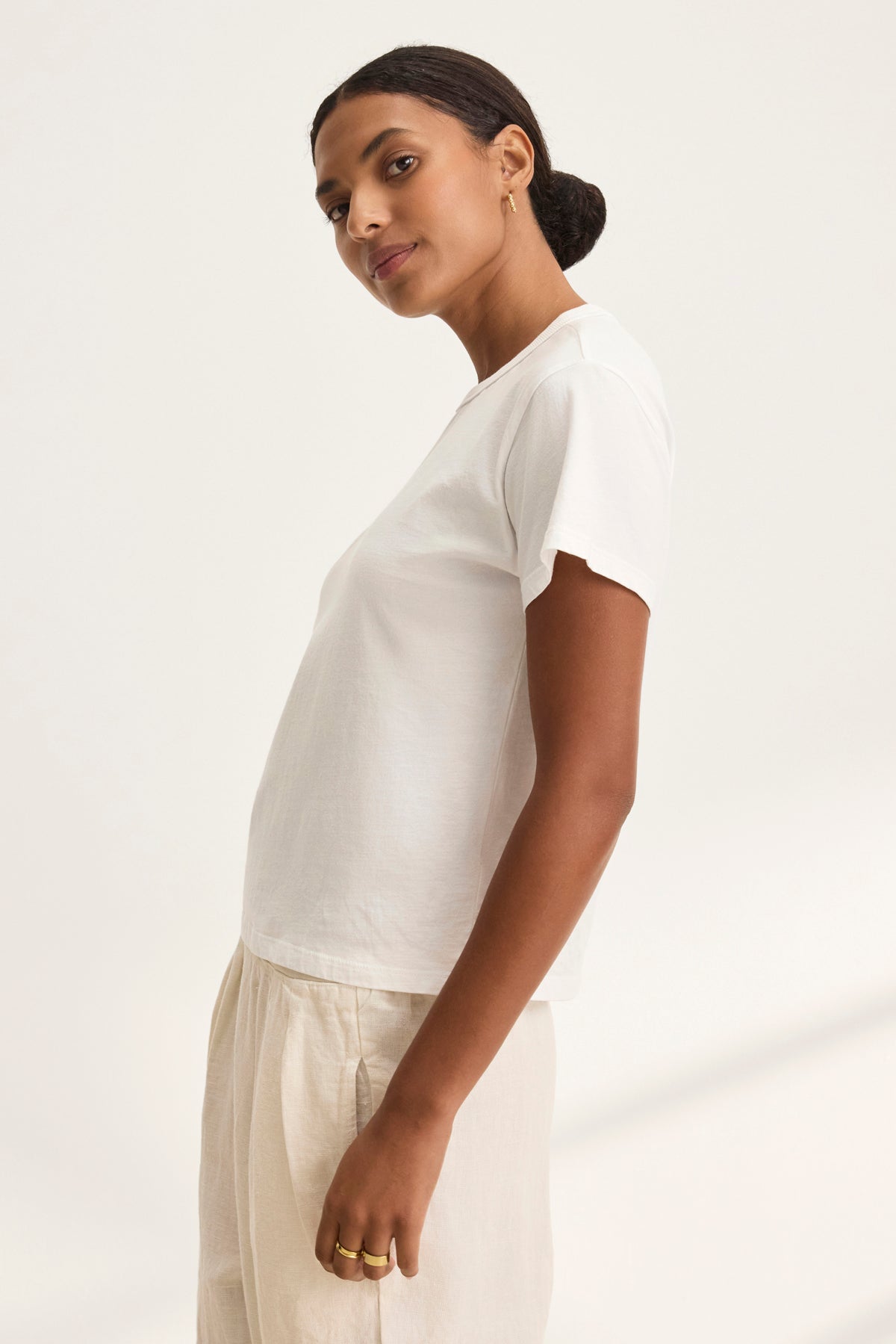   The individual is wearing a JAZZ TEE from Velvet by Graham & Spencer, featuring a relaxed boxy silhouette, paired with beige pants and standing sideways against a plain background. 