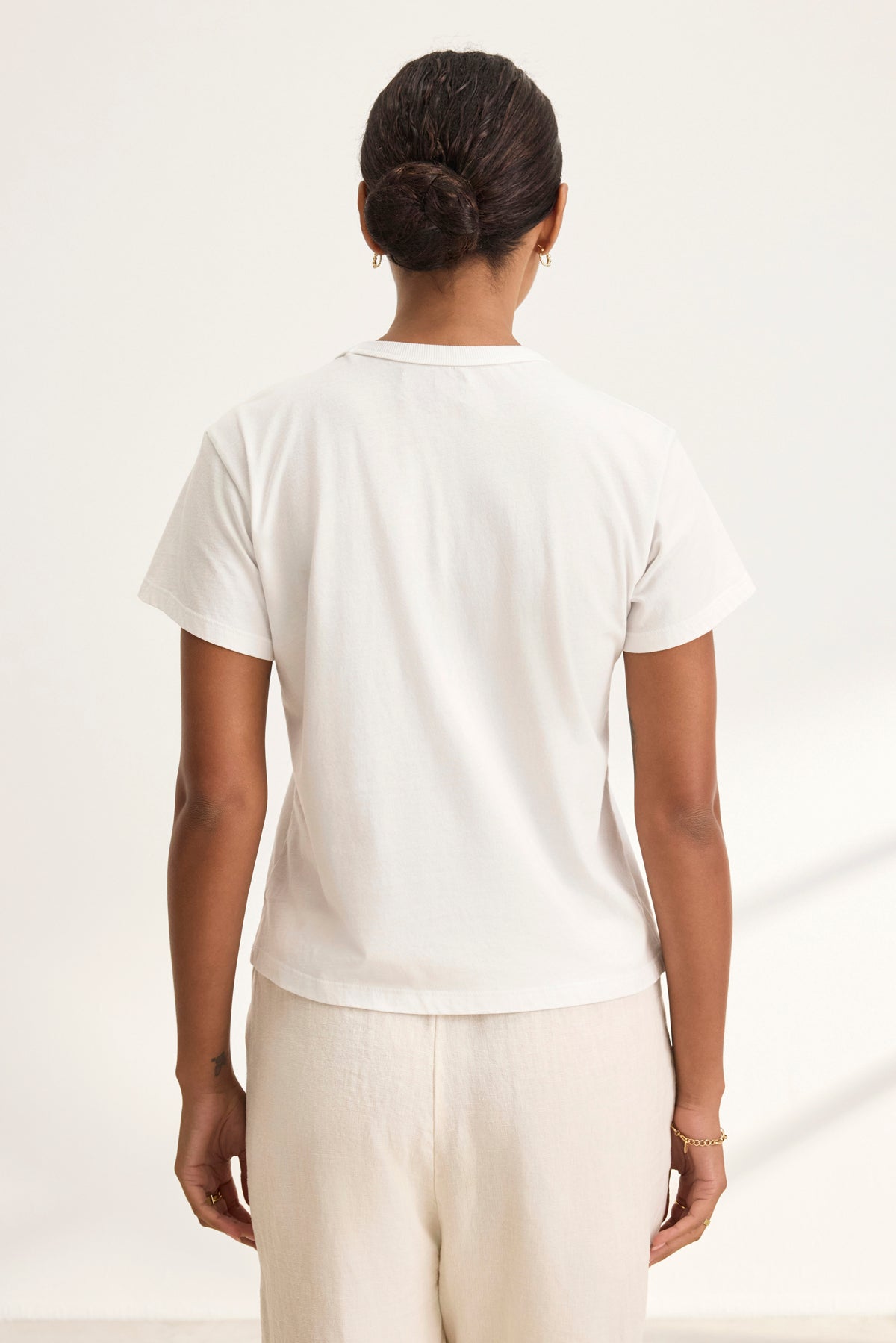   A dark-haired person with a bun is seen from behind wearing a Velvet by Graham & Spencer's JAZZ TEE in white organic cotton jersey, paired with cream pants. The shirt boasts a relaxed boxy silhouette and crew neckline, adding an effortless style against the neutral background. 