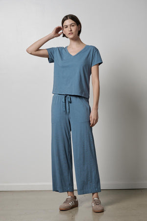 The model is wearing a blue PISMO PANT jumpsuit with slash pockets made of organic cotton by Velvet by Jenny Graham.