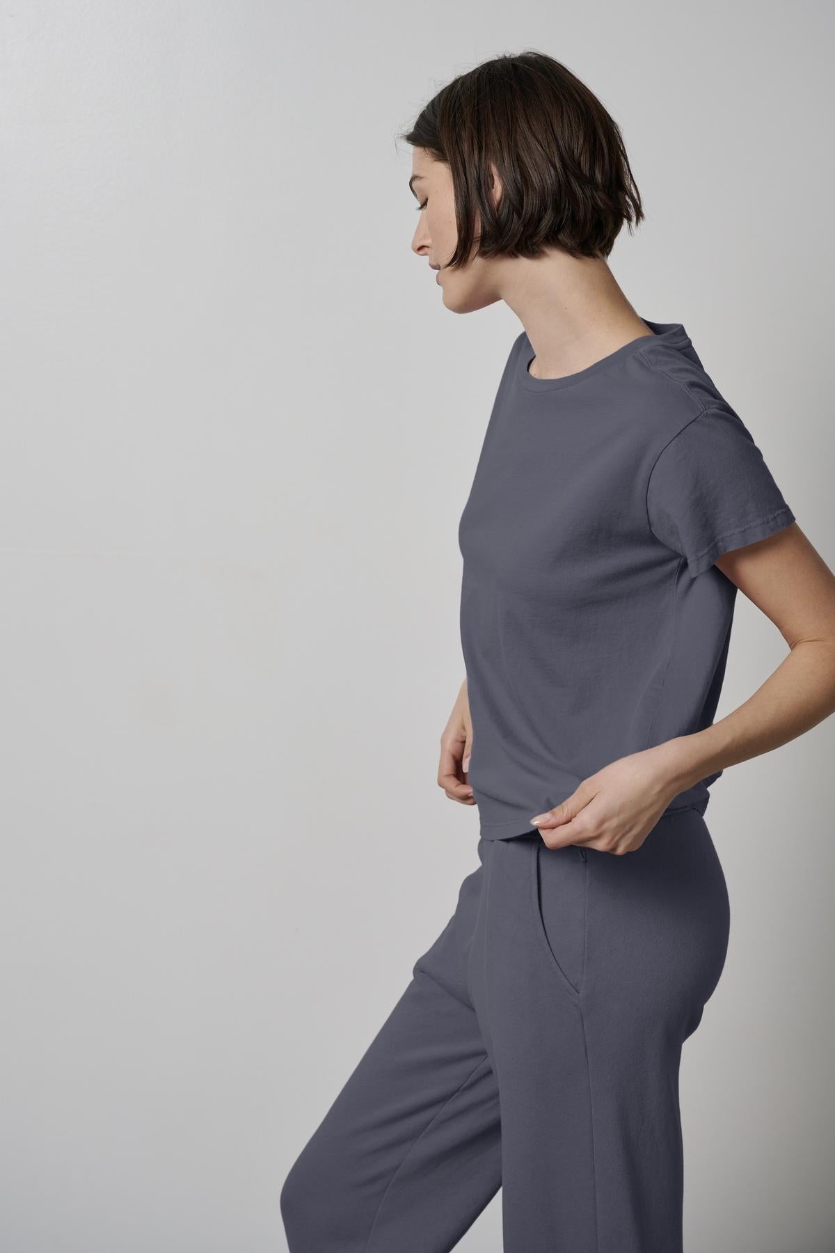   A person with short hair stands against a plain, light-colored wall, wearing a relaxed-fit TOPANGA TEE and pants made from eco-friendly materials by Velvet by Jenny Graham, looking down to their right. 