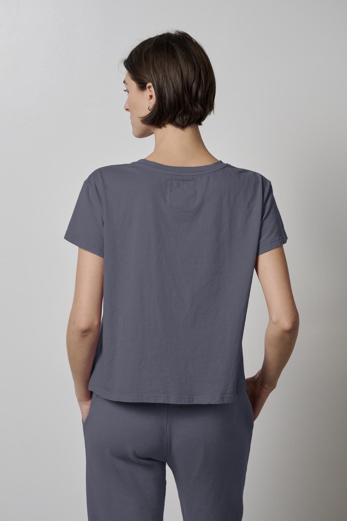   A person with short hair is standing with their back to the camera, wearing a Velvet by Jenny Graham TOPANGA TEE and organic cotton grey pants. 