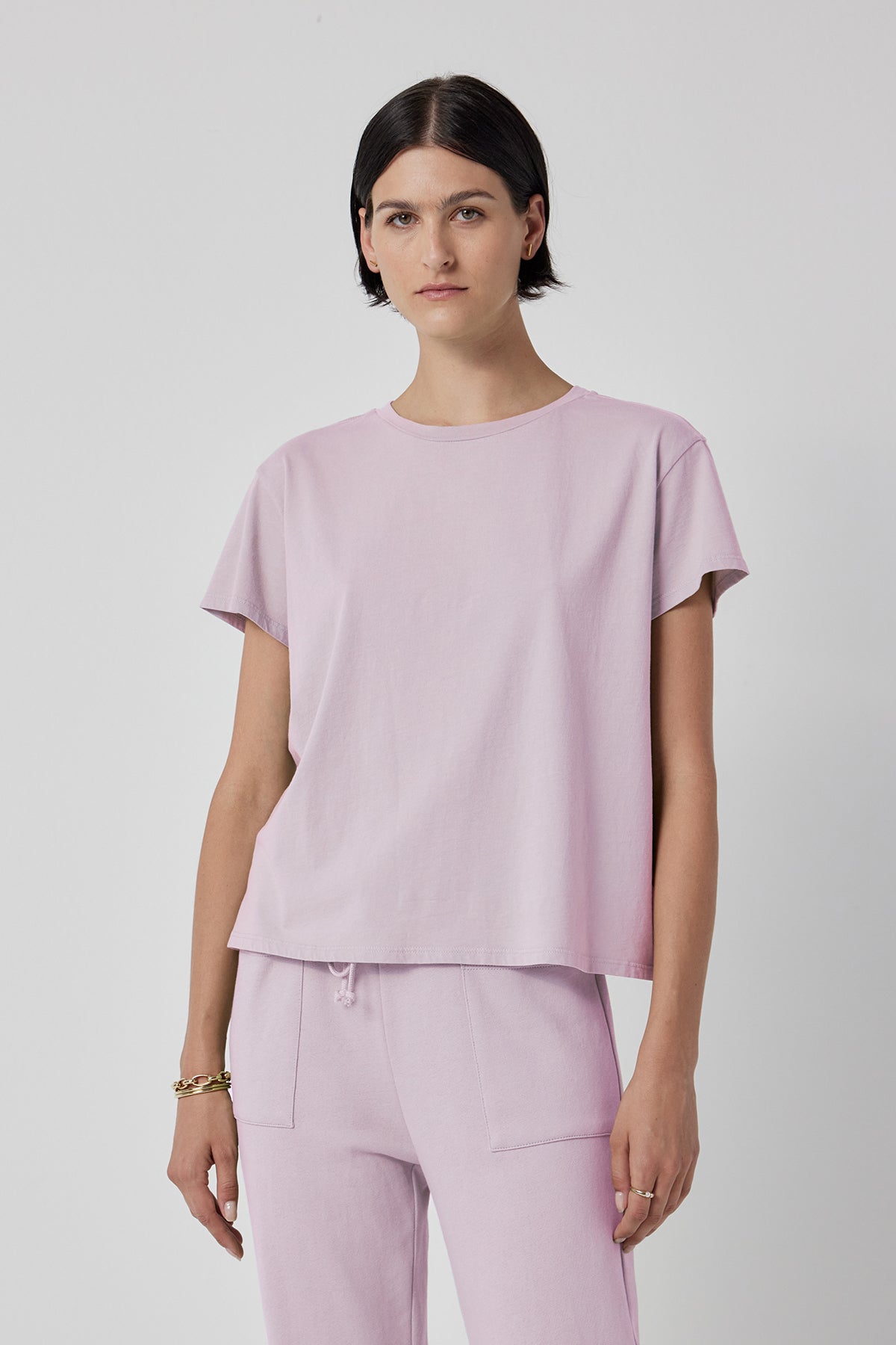   A person stands against a plain background wearing the TOPANGA TEE from Velvet by Jenny Graham, a light pink, relaxed-fit T-shirt paired with matching pants. The outfit, made of organic cotton, complements their short dark hair and neutral expression. 