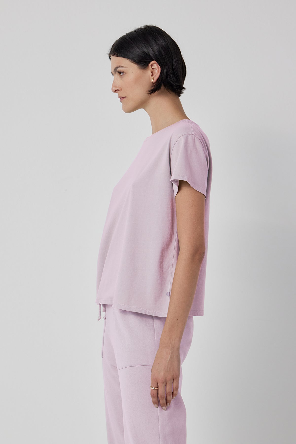 A person stands against a plain background, wearing the short-sleeved, light purple TOPANGA TEE from Velvet by Jenny Graham and matching organic cotton pants. They are looking to the left.-37862282297537