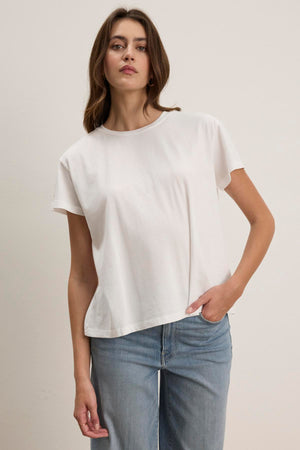 A person wearing the TOPANGA TEE from Velvet by Jenny Graham, featuring a relaxed fit in plain white organic cotton, paired with blue jeans, poses against a neutral background.