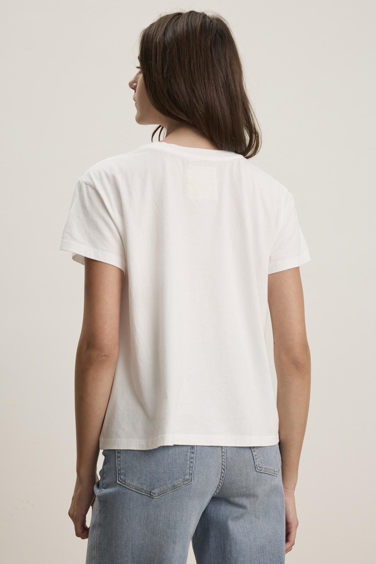   The individual is wearing the TOPANGA TEE by Velvet by Jenny Graham, which is a relaxed-fit white T-shirt made from organic cotton, paired with blue jeans and facing away. 