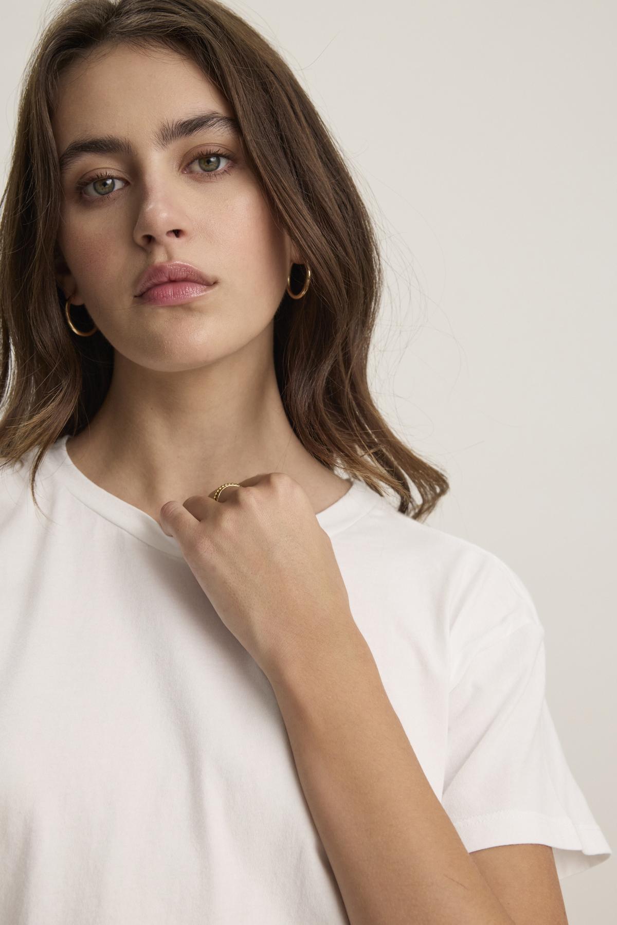   A person with long hair and hoop earrings wears the TOPANGA TEE, a relaxed-fit, organic cotton white t-shirt from Velvet by Jenny Graham. They are looking forward with a neutral expression and have their hand near their chin. 