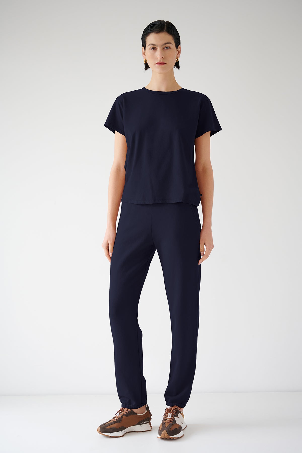   A woman in a navy blue organic cotton t-shirt and ZUMA SWEATPANT stands against a white background, wearing brown and beige sneakers by Velvet by Jenny Graham. 