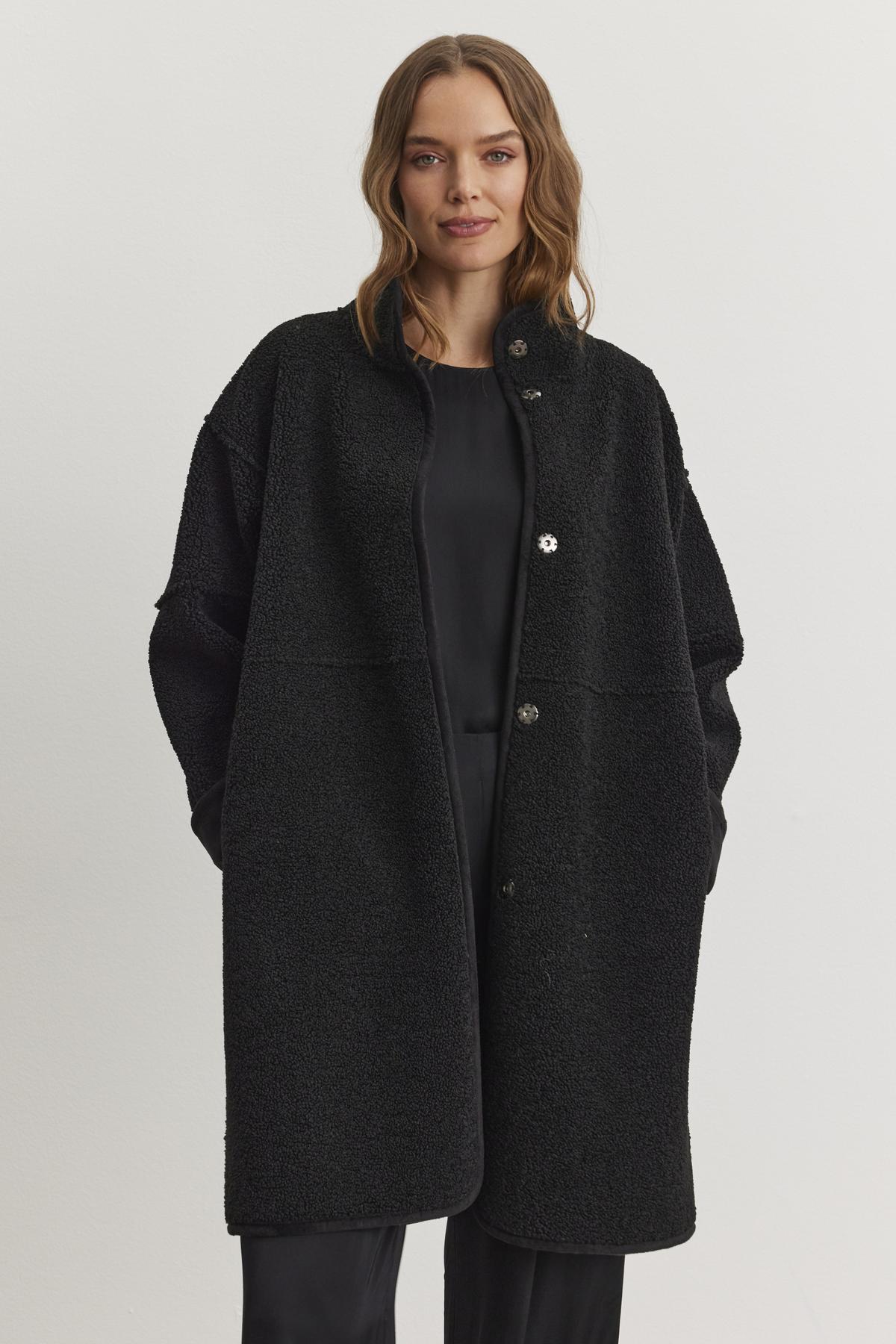   A person stands against a plain light background, wearing the CARA LUXE SHERPA REVERSIBLE JACKET from Velvet by Graham & Spencer—a luxurious reversible coat with sherpa detailing—over a sleek black outfit. 