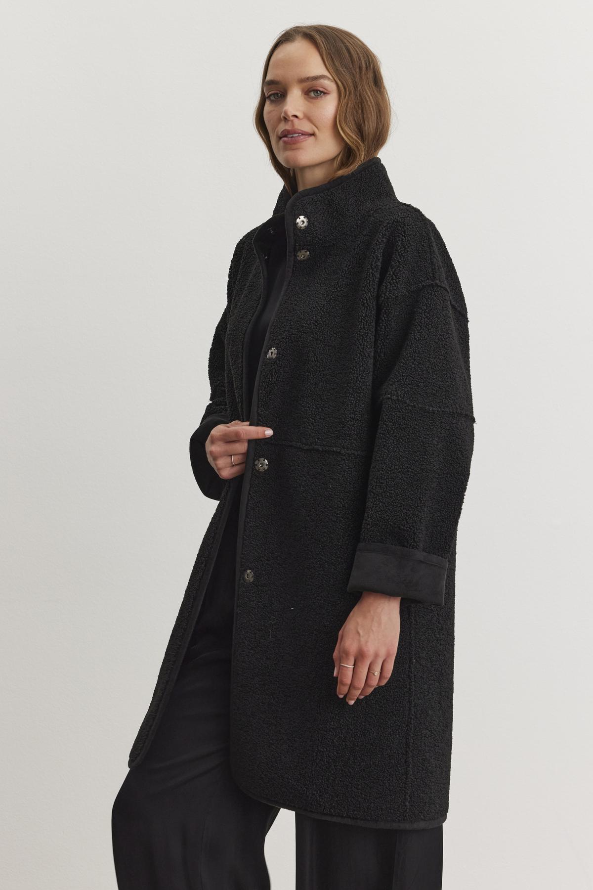 A person wearing a black Cara Luxe Sherpa Reversible Jacket by Velvet by Graham & Spencer, featuring a faux suede texture, stands against a plain background, looking forward.-38219221926081