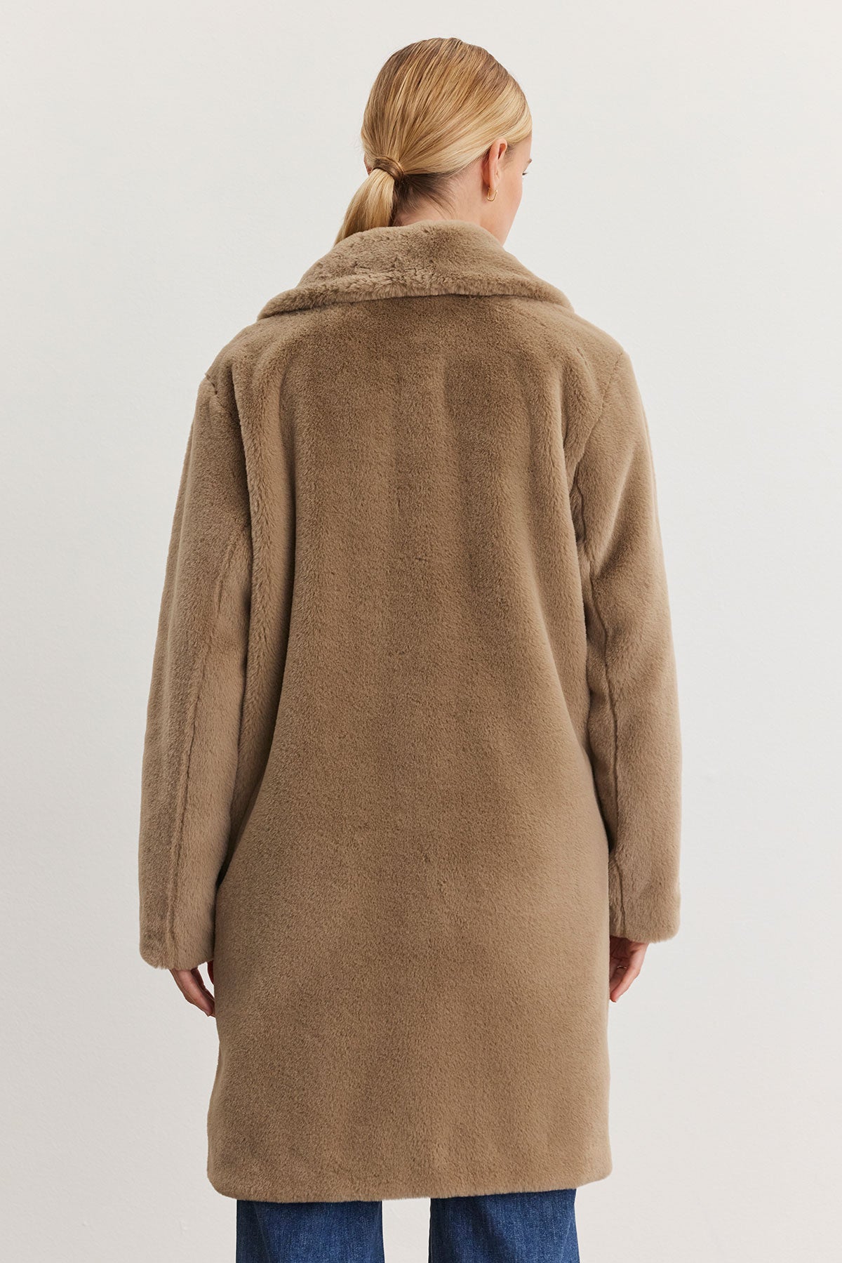   A person with blonde hair styled in a bun wears the EVALYN LUX FAUX FUR COAT by Velvet by Graham & Spencer; its long, brown design is perfect for a seamless transition from day to night, as they face away. 