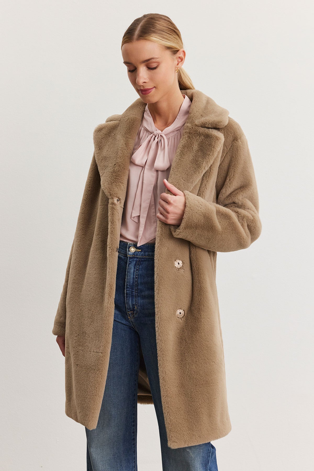   The individual, wrapped in the EVALYN LUX FAUX FUR COAT from Velvet by Graham & Spencer, exudes day-to-night elegance against a simple backdrop, paired with a light pink blouse and blue jeans. 