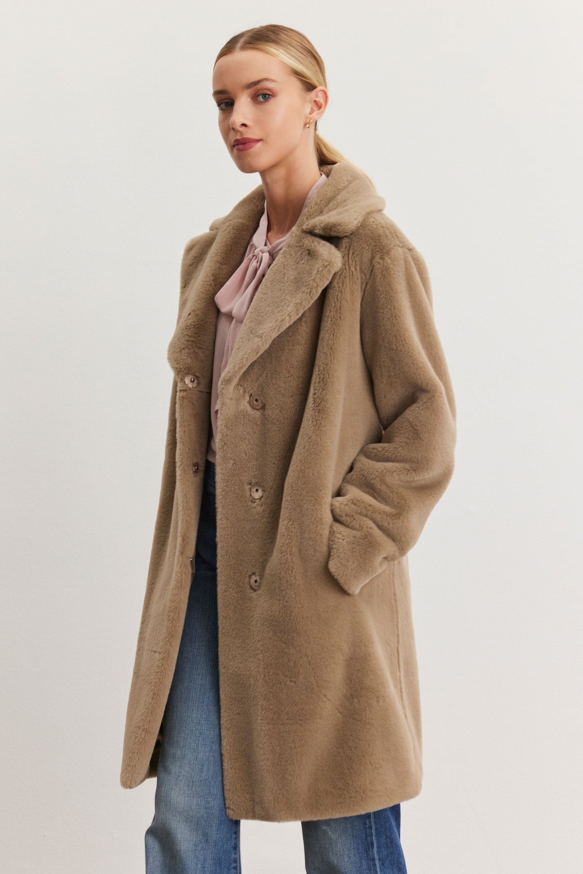   A woman wearing the EVALYN LUX FAUX FUR COAT by Velvet by Graham & Spencer, a camel-hued bundle-up coat made of faux fur, stands gracefully against a neutral backdrop. Her ensemble effortlessly transitions from day to night with a chic pink blouse and blue jeans. 