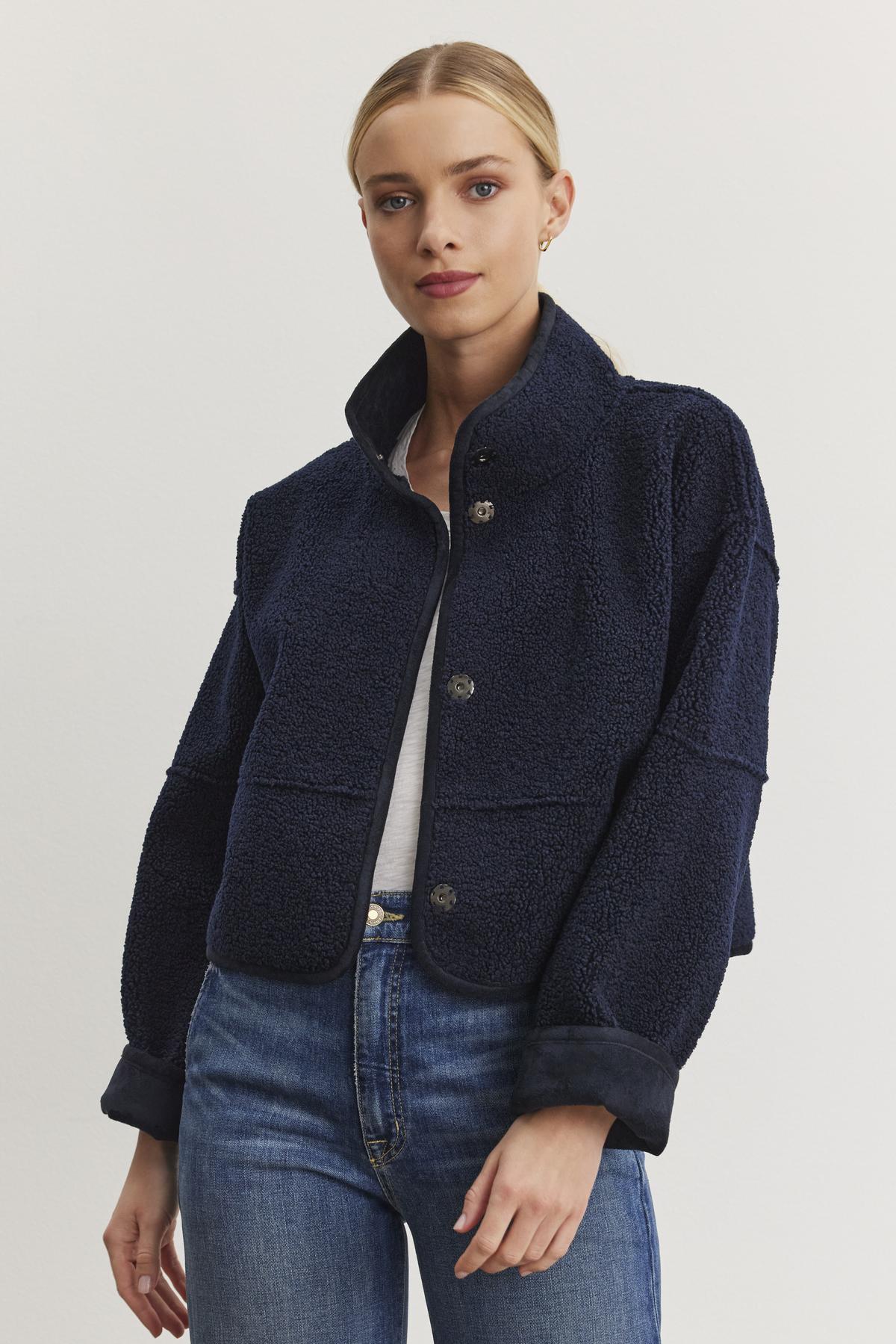   A woman poses against a plain white backdrop, showcasing the Velvet by Graham & Spencer's KELLY LUXE SHERPA REVERSIBLE JACKET in a chic navy blue wool with faux sherpa detailing, layered over a white shirt and blue jeans. 