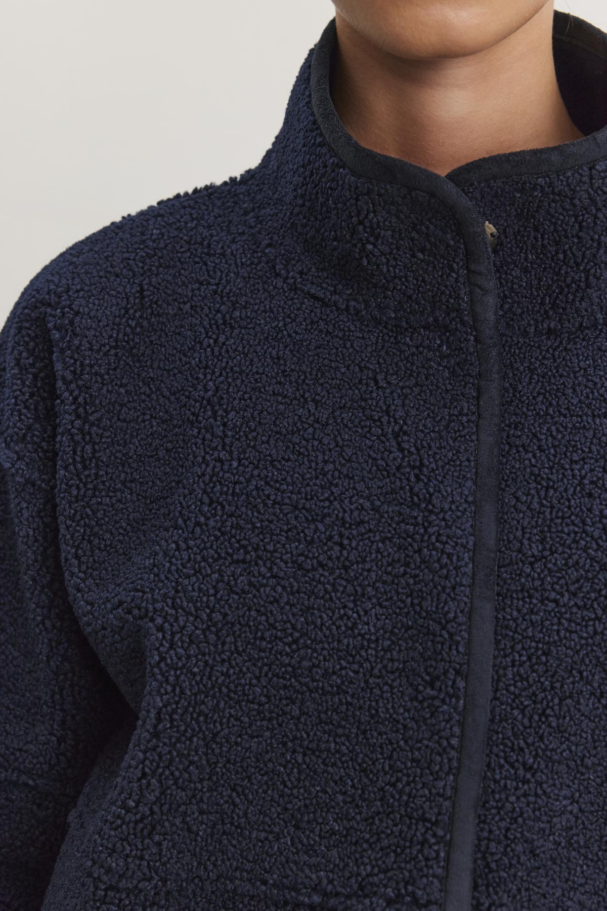   A close-up of an individual wearing the Velvet by Graham & Spencer KELLY LUXE SHERPA REVERSIBLE JACKET, a dark blue textured fleece coat with a high collar and faux sherpa lining for enhanced warmth and comfort. 