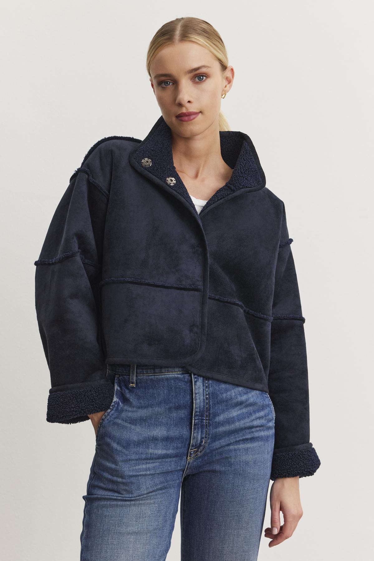   Dressed in a dark blue KELLY LUXE SHERPA REVERSIBLE JACKET from Velvet by Graham & Spencer over a white top and blue jeans, the person stands against a plain background, radiating effortless style. 