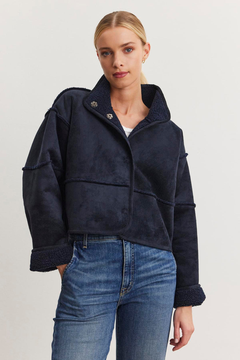 Dressed in a dark blue KELLY LUXE SHERPA REVERSIBLE JACKET from Velvet by Graham & Spencer over a white top and blue jeans, the person stands against a plain background, radiating effortless style.