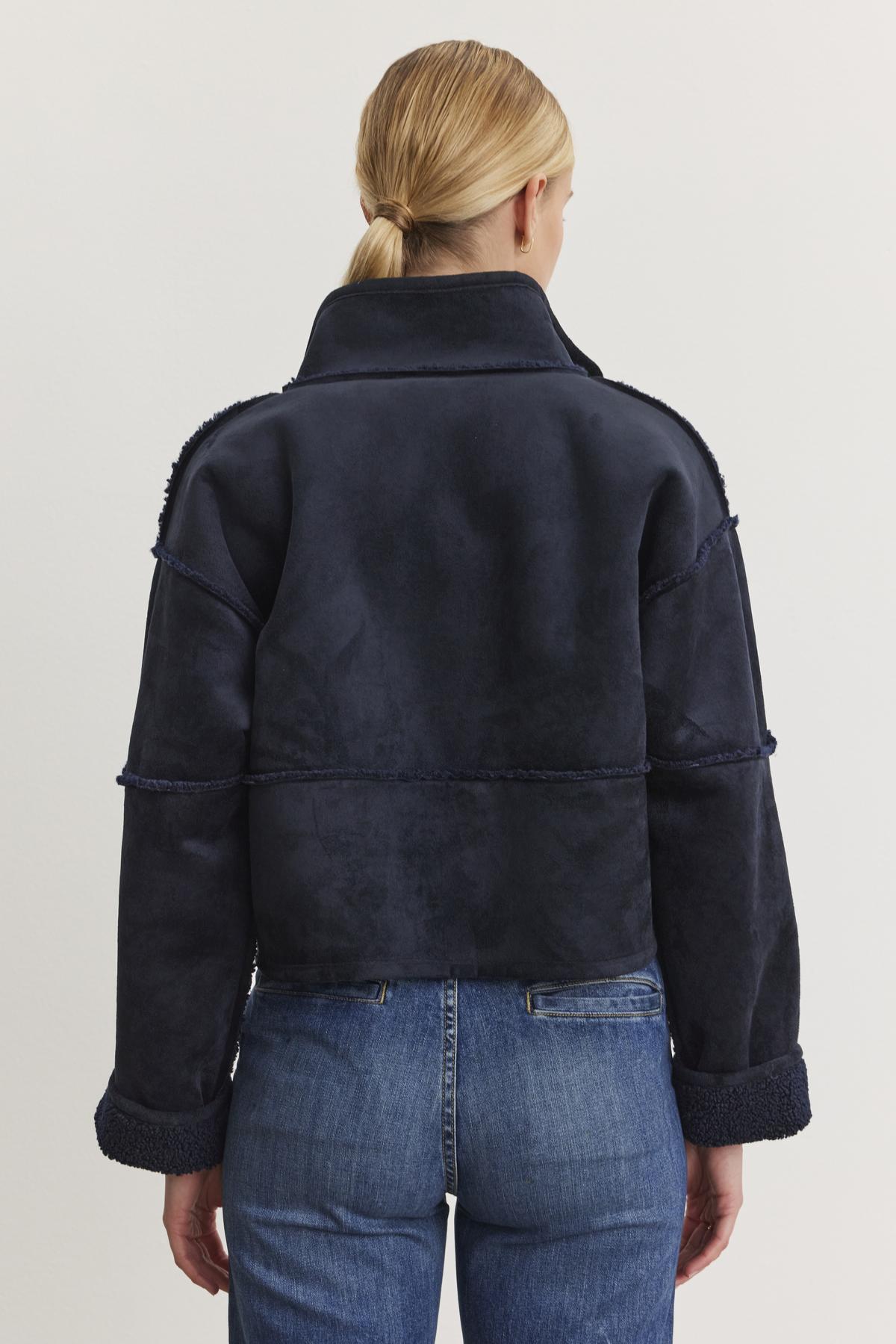 A person with tied-back hair wears the KELLY LUXE SHERPA REVERSIBLE JACKET by Velvet by Graham & Spencer, featuring a high collar and paired with blue jeans, standing with their back to the camera.-38219210096833