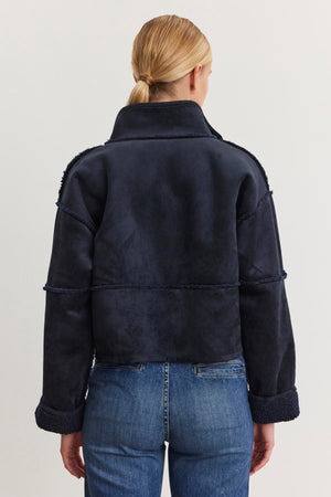 A person with tied-back hair wears the KELLY LUXE SHERPA REVERSIBLE JACKET by Velvet by Graham & Spencer, featuring a high collar and paired with blue jeans, standing with their back to the camera.