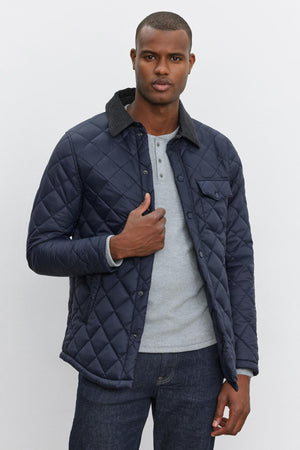 A person in a LOU JACKET by Velvet by Graham & Spencer, featuring a quilted puffer design with a corduroy collar, is wearing a light grey henley shirt and blue jeans.