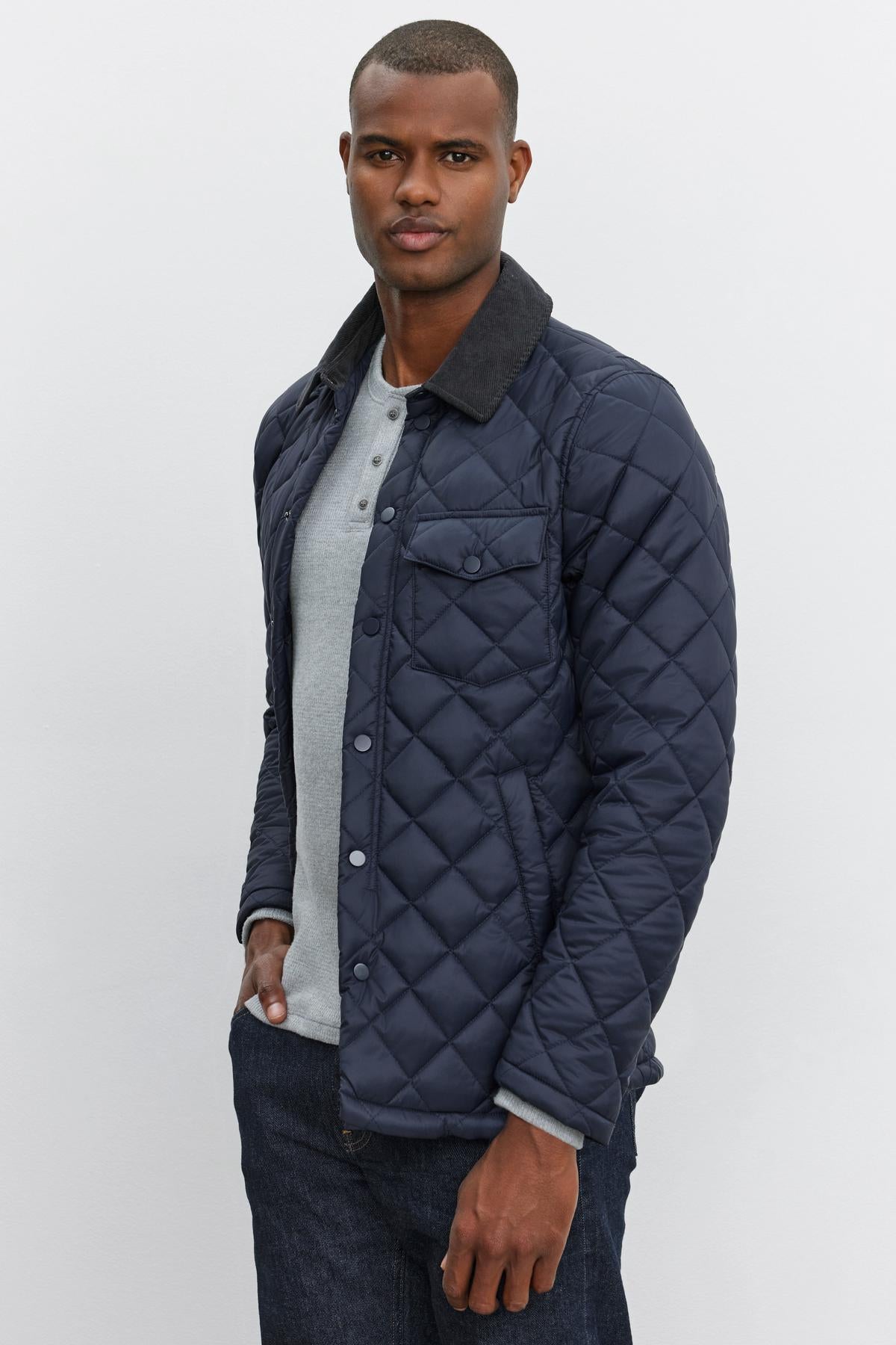   A man wearing the LOU JACKET by Velvet by Graham & Spencer, a dark blue quilted puffer jacket with a corduroy collar, over a light gray henley shirt and dark jeans, stands in front of a plain white background. 