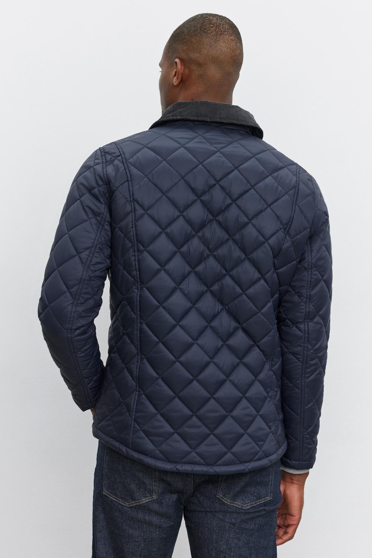   A man is wearing the Velvet by Graham & Spencer LOU JACKET, a dark blue quilted puffer with a corduroy collar, viewed from the back. He has short hair and is also sporting dark blue jeans. 