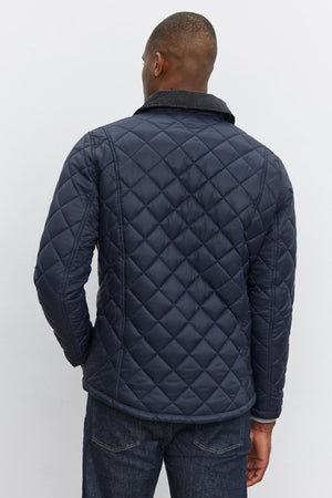 A man is wearing the Velvet by Graham & Spencer LOU JACKET, a dark blue quilted puffer with a corduroy collar, viewed from the back. He has short hair and is also sporting dark blue jeans.