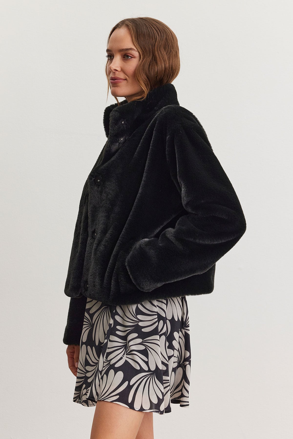   A person modeling the SHEENA LUXE FAUX FUR JACKET by Velvet by Graham & Spencer, featuring a cozy fit and mock neck collar in black, stands against a white background, complemented by a patterned skirt. 